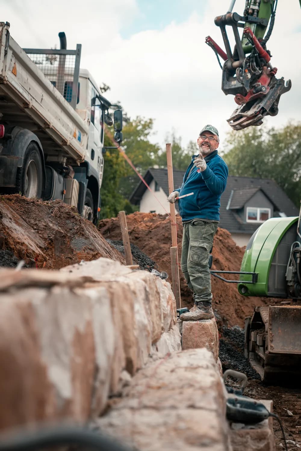 Facharbeiter beim Bau einer Natursteinmauer auf der Baustelle, im Hintergrund Kipper und Bagger