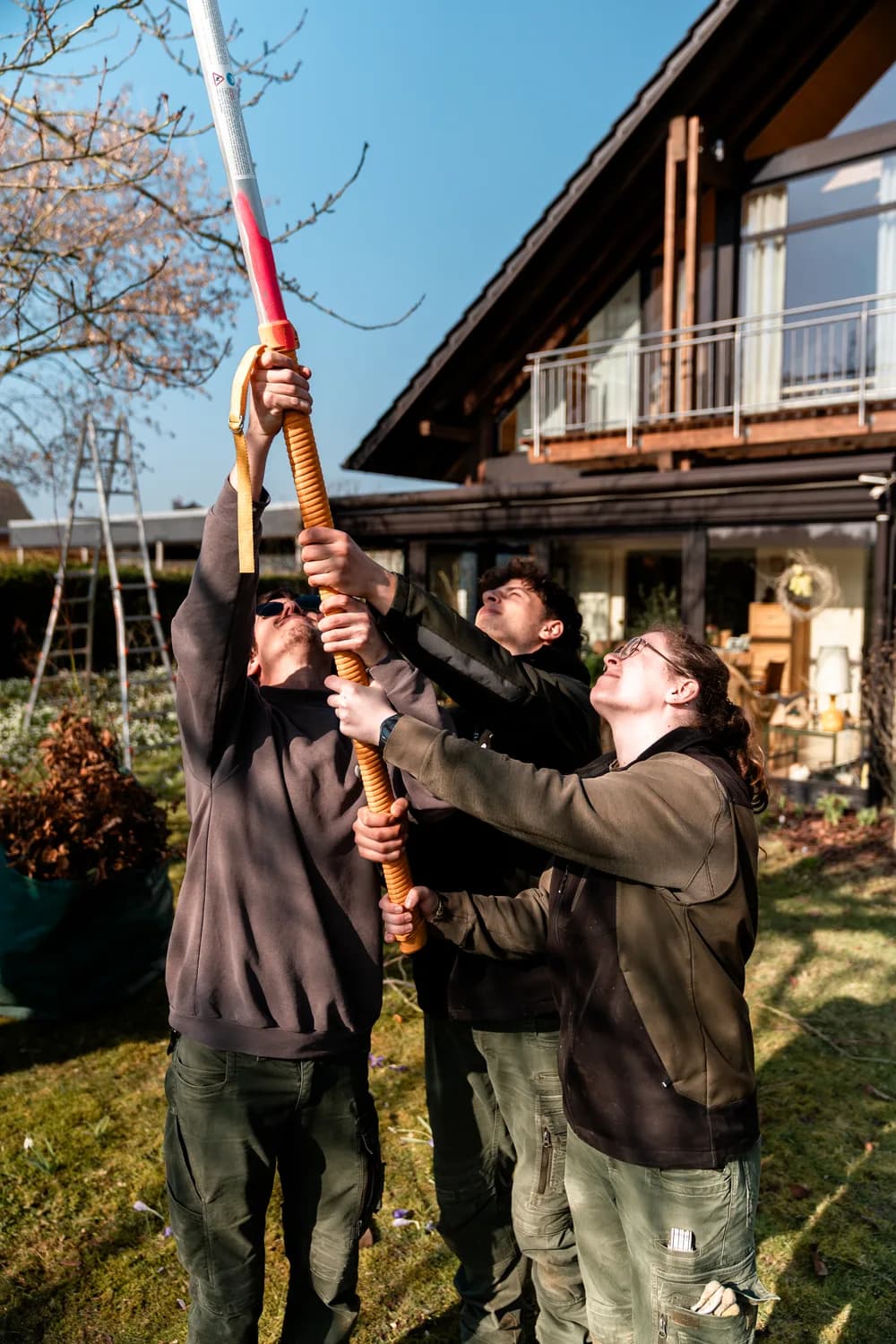 Drei junge Teammitglieder der Scheel-Gärten hieven gemeinsam eine lange Teleskop-Astsäge nach oben und packen dabei zusammen an. Echtes Foto.