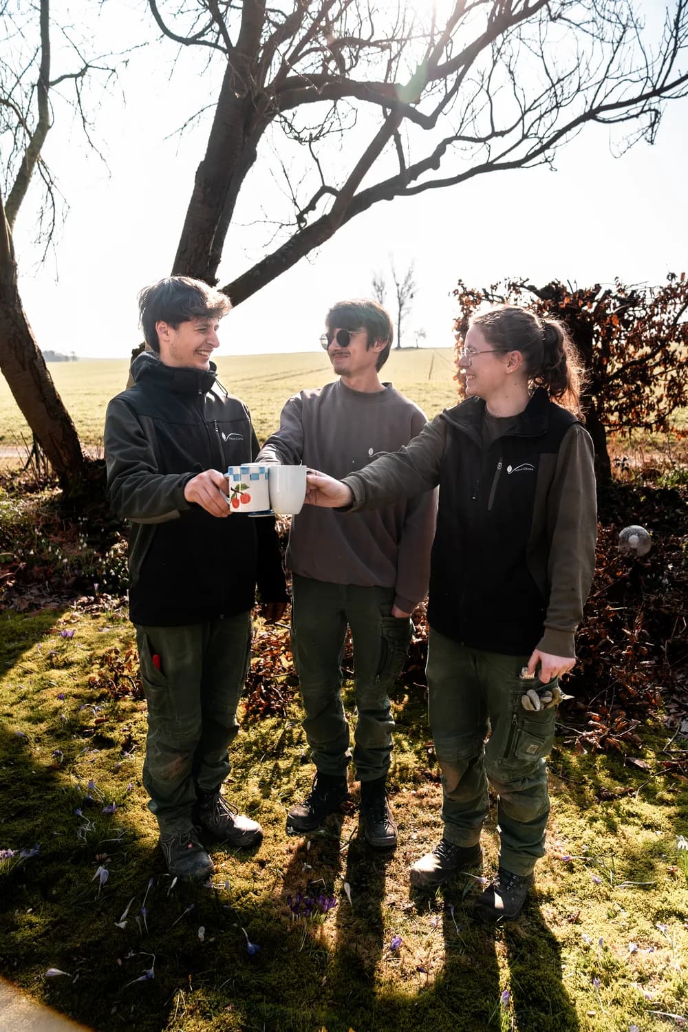 Drei junge Kolleginnen und Kollegen der Scheel-Gärten stoßen auf einer Wiese mit Kaffeebechern an und lachen zusammen. Echtes Foto.